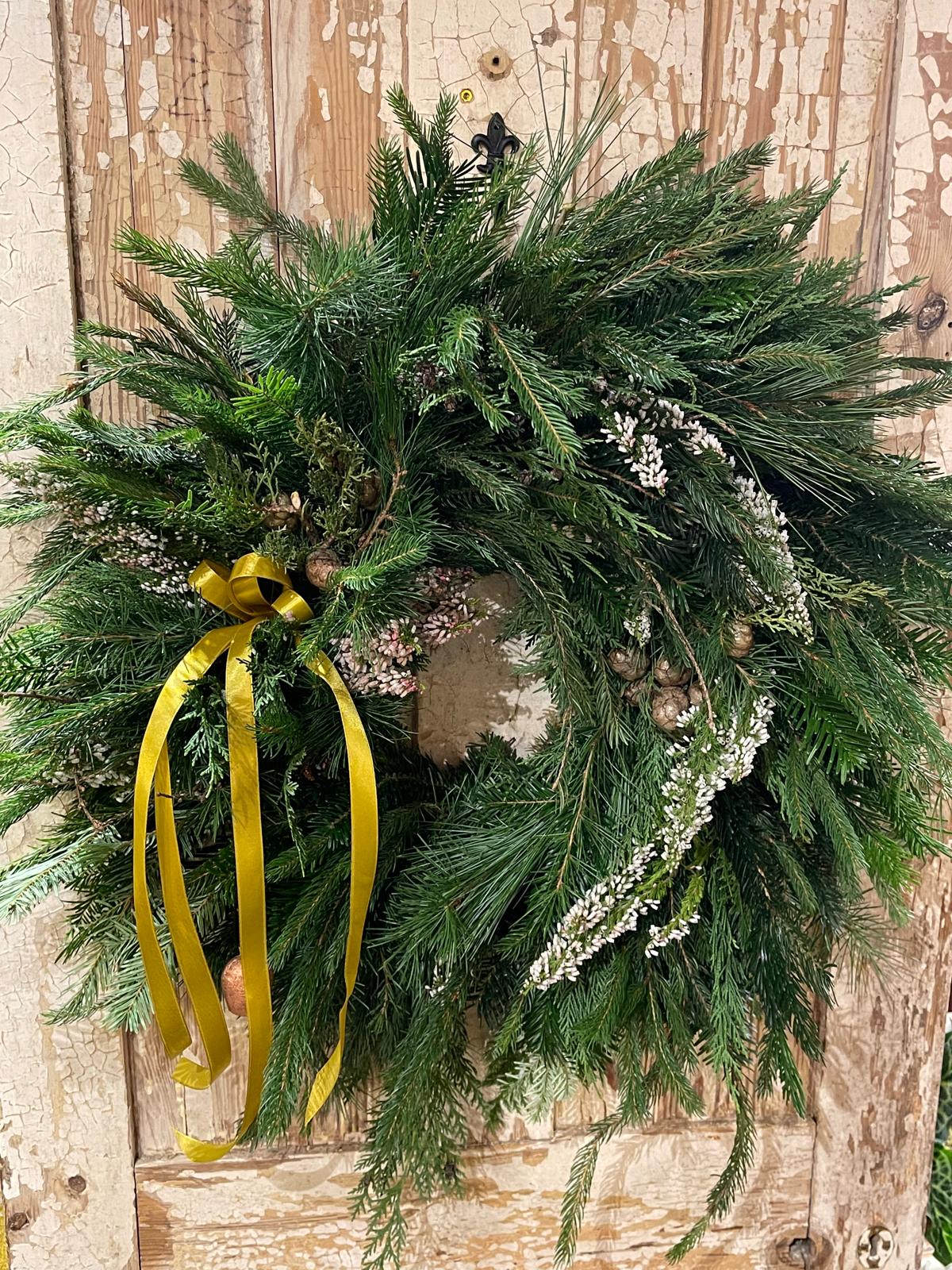 A rustic green pine wreath with a gold ribbon bow, hanging against a distressed wooden door. The wreath is decorated with small pinecones and white berries.