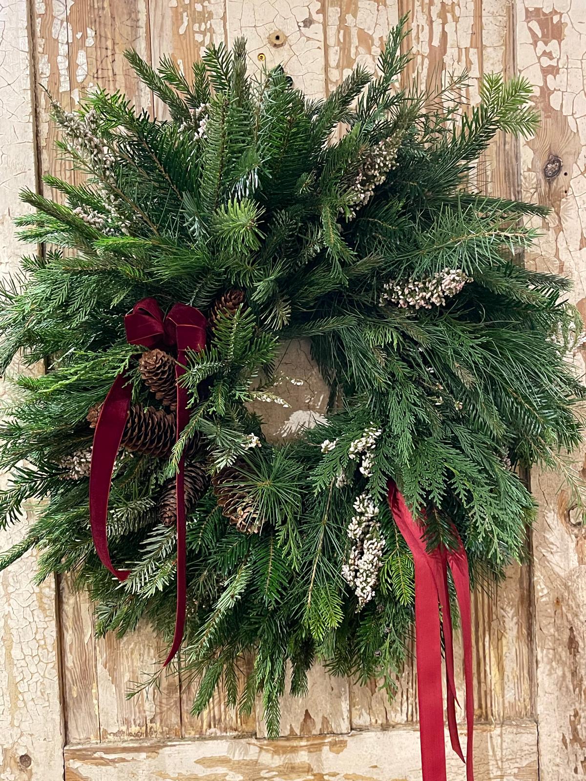 A lush green pine wreath with pinecones and white berries, decorated with a deep red velvet ribbon bow and hanging ribbons, displayed against a rustic, weathered wooden door.
