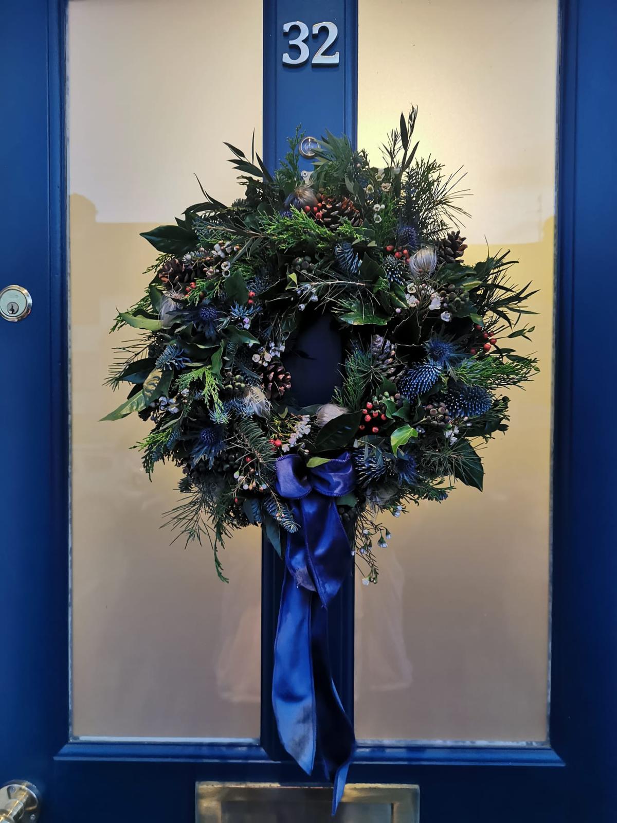 A festive Christmas wreath with green pine and fir branches, blue berries, pinecones, and a large blue satin bow, hanging on a blue front door with the number 32 visible.