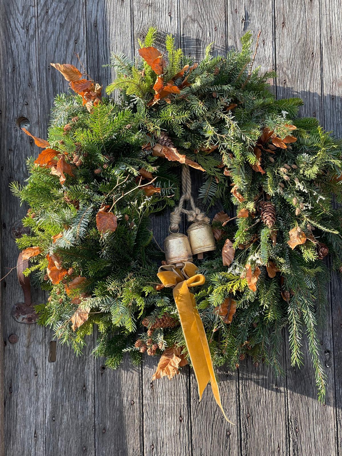 A rustic holiday wreath made of green pine and fir branches, decorated with dried orange-brown leaves, small pinecones, two hanging metal bells, and a long mustard-yellow ribbon, displayed against a weathered gray wooden wall.