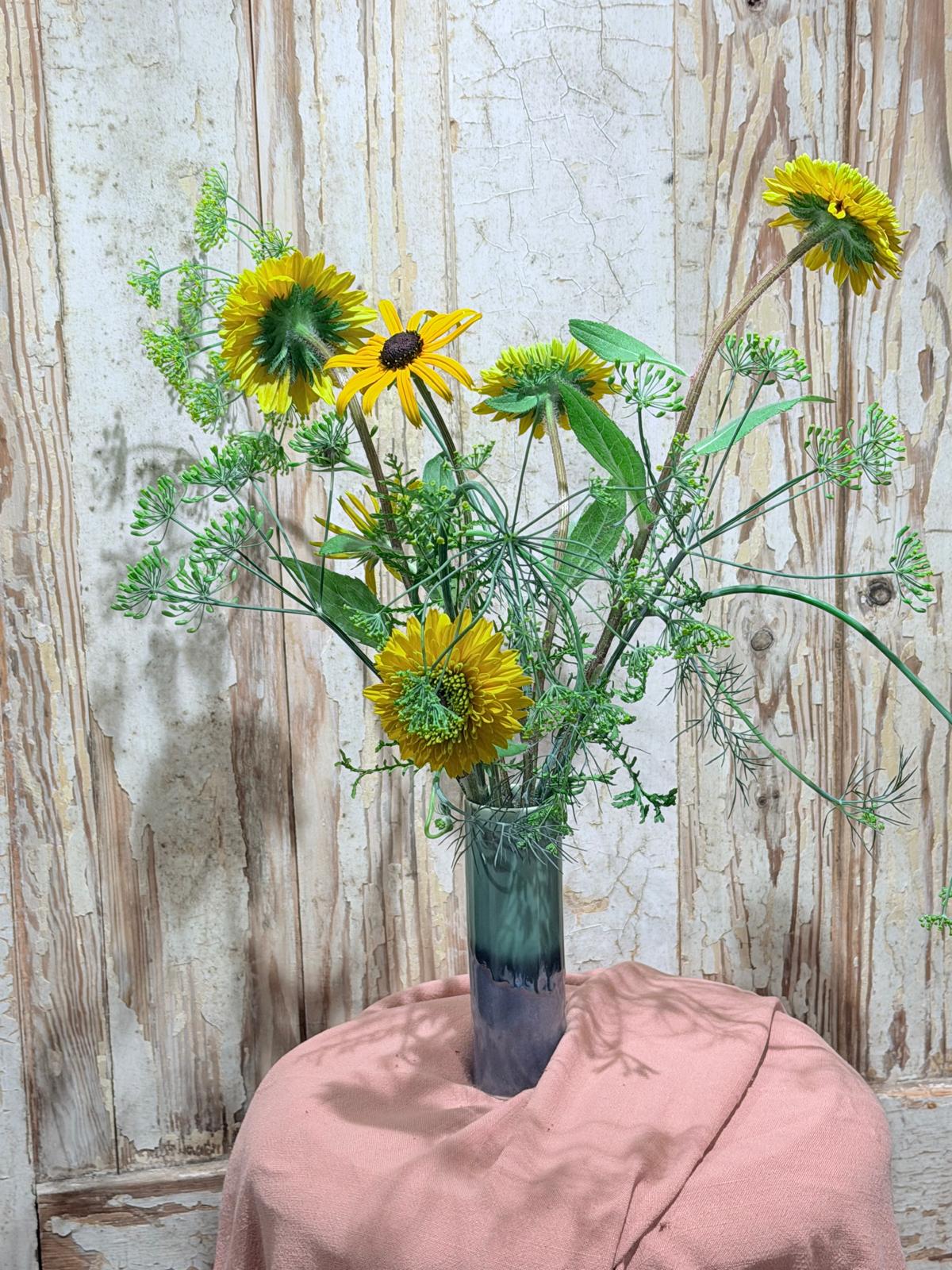 A floral arrangement of yellow flowers and green foliage in a dark blue-green glass vase, set against a rustic whitewashed wooden wall with a draped dusty pink cloth underneath.