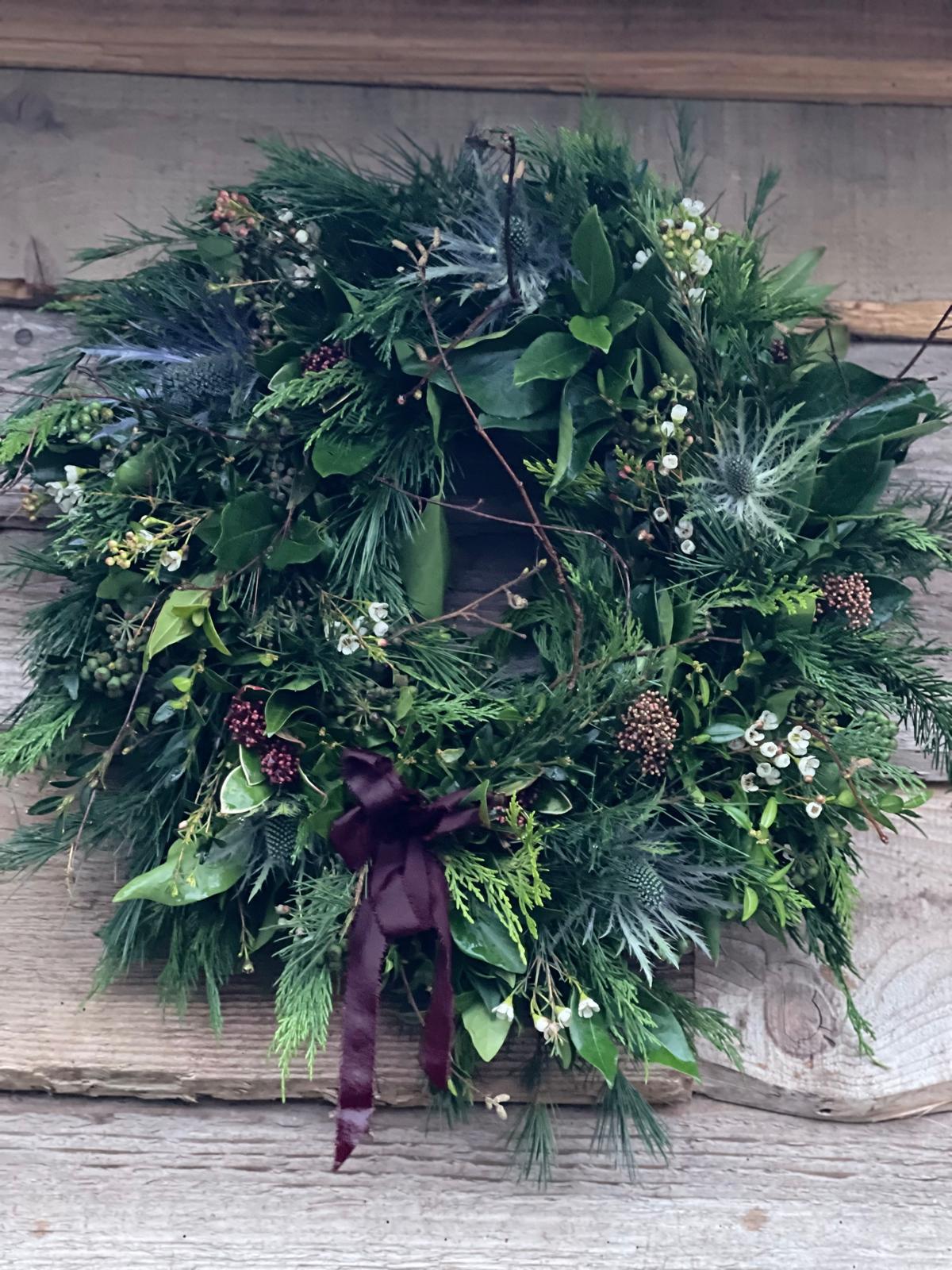 A lush green holiday wreath with pine branches, evergreen foliage, small white berries, and a dark burgundy ribbon bow, displayed against a rustic wooden plank background.